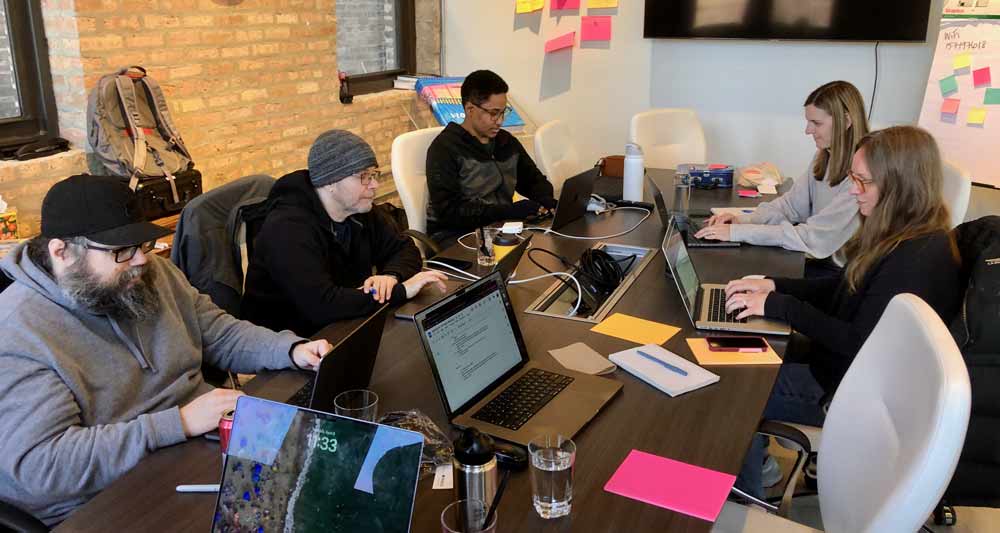 Image of five people working at laptops around a table.